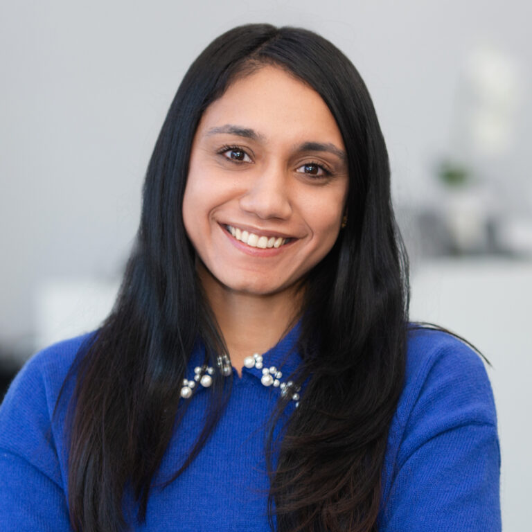 A woman with long black hair, wearing a blue sweater with a pearl necklace detail, smiles at the camera in a softly lit indoor setting.