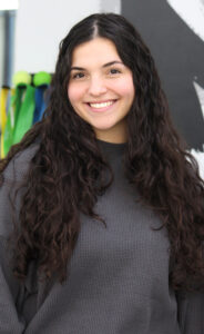 A woman with long, curly dark hair and a gray sweater smiles at the camera, standing indoors with colorful ribbons and a black-and-white wall decoration in the background.