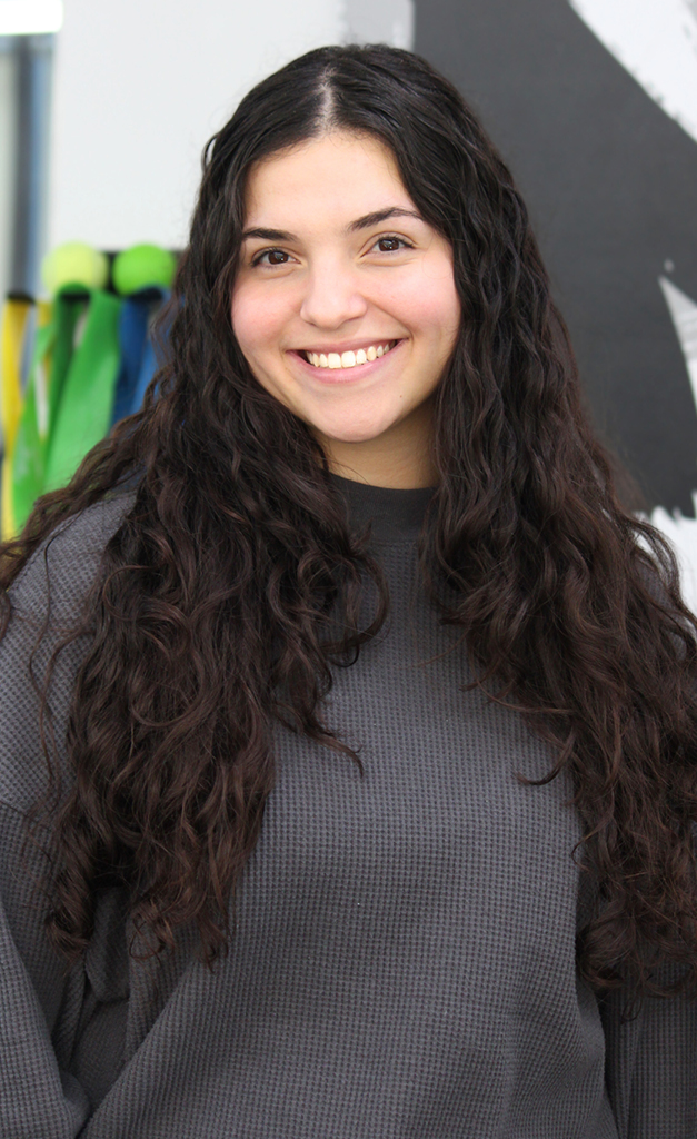 A woman with long, curly dark hair and a gray sweater smiles at the camera, standing indoors with colorful ribbons and a black-and-white wall decoration in the background.