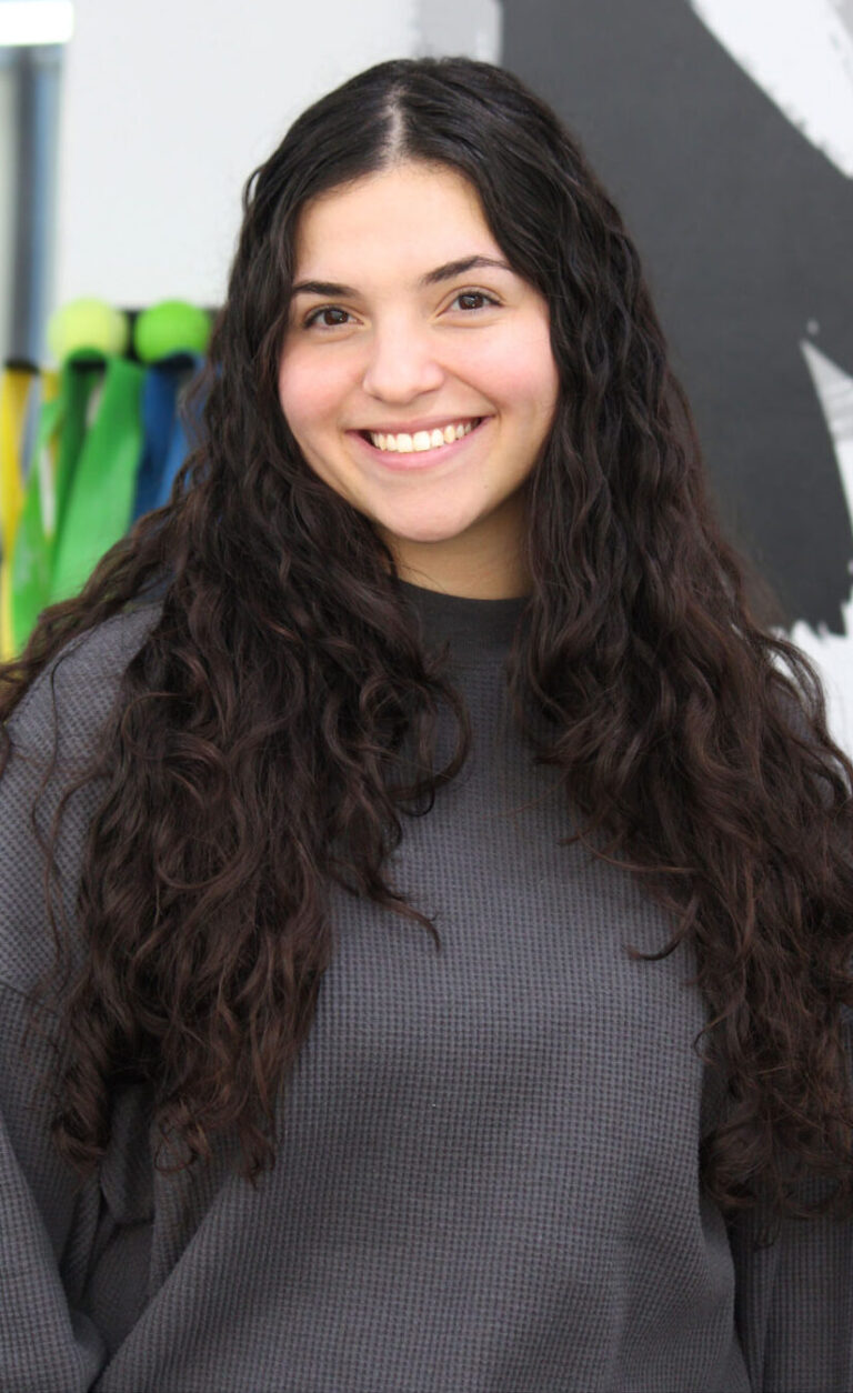 A young woman with long, curly dark hair smiles at the camera. She is wearing a loose gray sweater and standing indoors in front of a black-and-white abstract wall and some colorful objects in the background.
